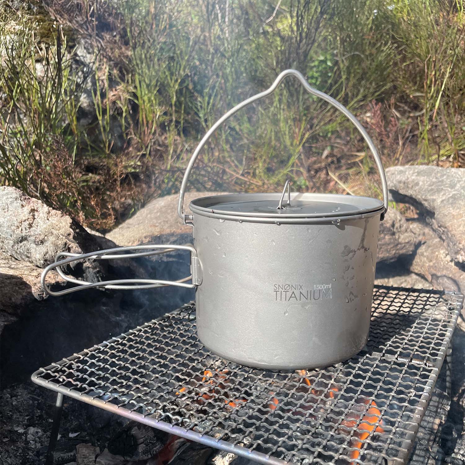 Titan turkjele på 1500 ml plassert over et bål på en rist, omgitt av natur og røyk fra bålet.