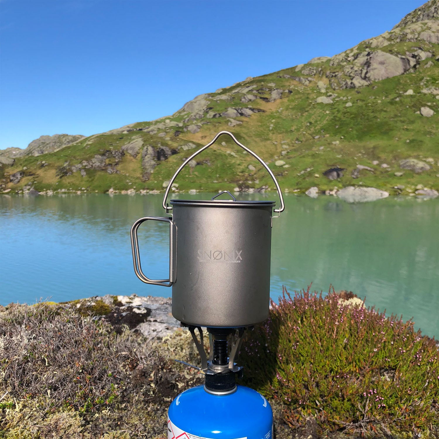 En 700 ml titan turkjele festet til en gassbrenner ved et naturskjønt fjellvann under blå himmel.